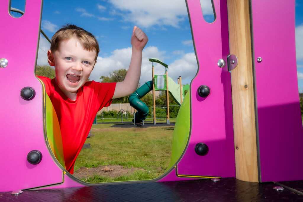A boy looking through a purple piece of play equipment with his hand in the air